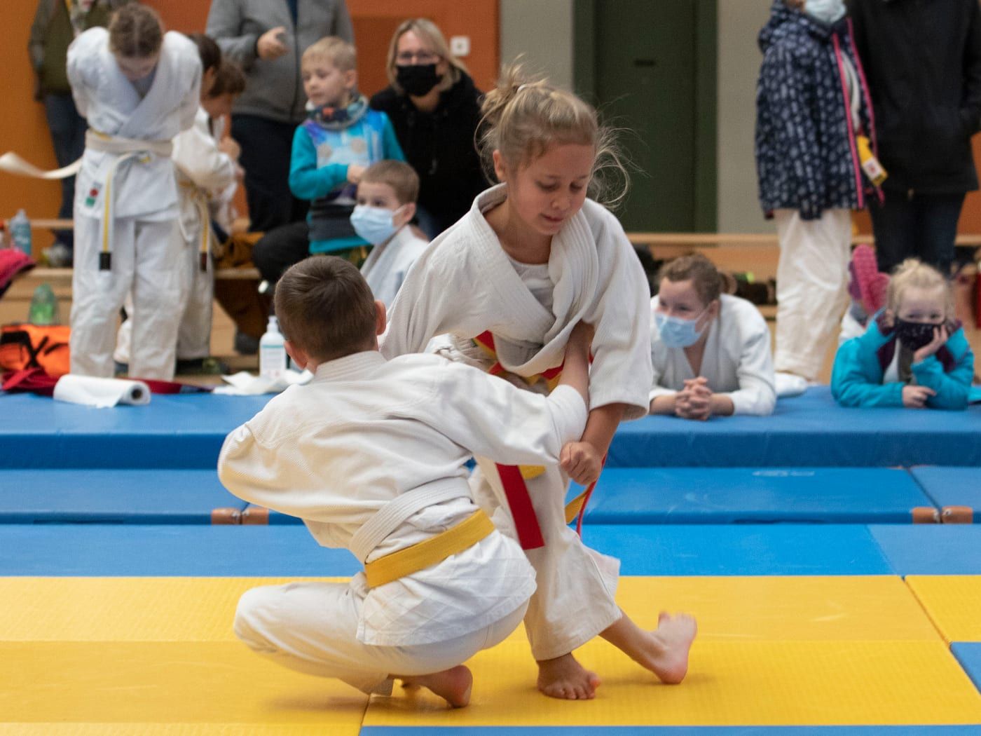 Ein Mädchen mit rotem Zusatzgürtel und ein Knabe mit gelbem Gürtel im Kampf auf blau-gelben Matten, dahinter schauen Kinder und Eltern zu.