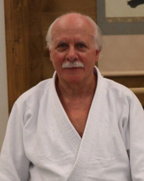 Älterer Mann mit weissem Haarkranz und weissem Schnurrbart in weissem Keikogi vor einer Holzwand.