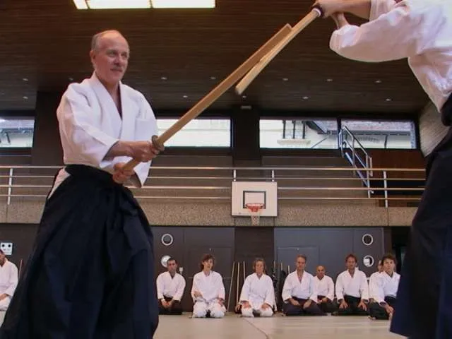 Ein Aikidoka in Hakama hält ein Holzschwert gegen das Schwert seines Partners, am Rand der Turnhalle knien Zuschauende in weissen Keikogi.