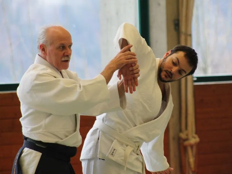 Ein älterer Aikidoka hält das abgewinkelte Handgelenk seines jungen Partners mit weissem Gürtel, der sich zur Seite neigt.