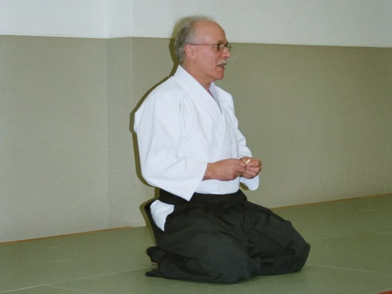 Ein Mann mit Brille in weissem Keikogi und schwarzem Hakama kniet auf grünen Matten und spricht.