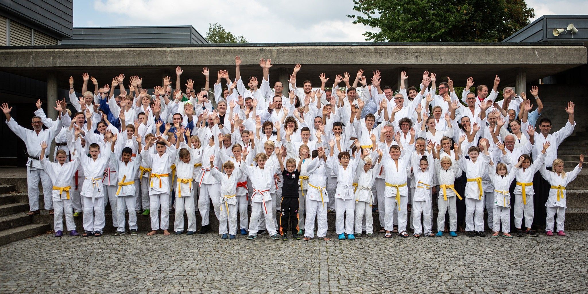 Gruppenfoto am OpenAir-Spektakel: über hundert Judoka und Aikidoka in weissen Anzügen und bunten Gürteln winken in die Kamera, vorne die Kinder.