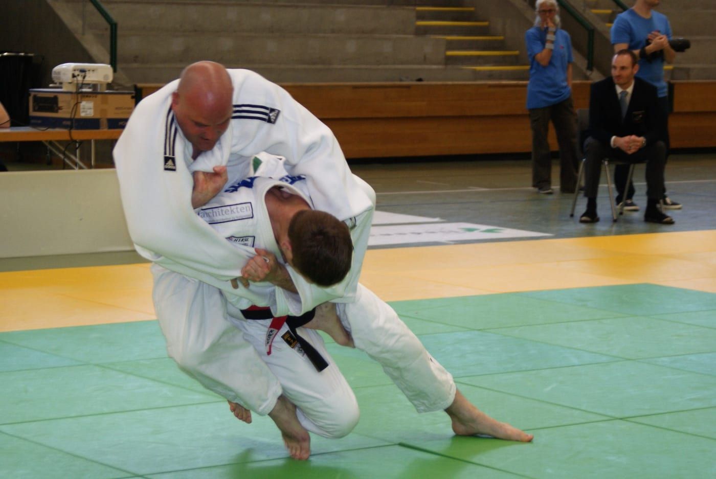 Zwei erwachsene Judokas im Wettkampf: Einer, mit schwarzem und rotem Gürtel, dreht zu einem Wurf ein. Am Mattenrand sitzt ein Kampfrichter im Anzug.