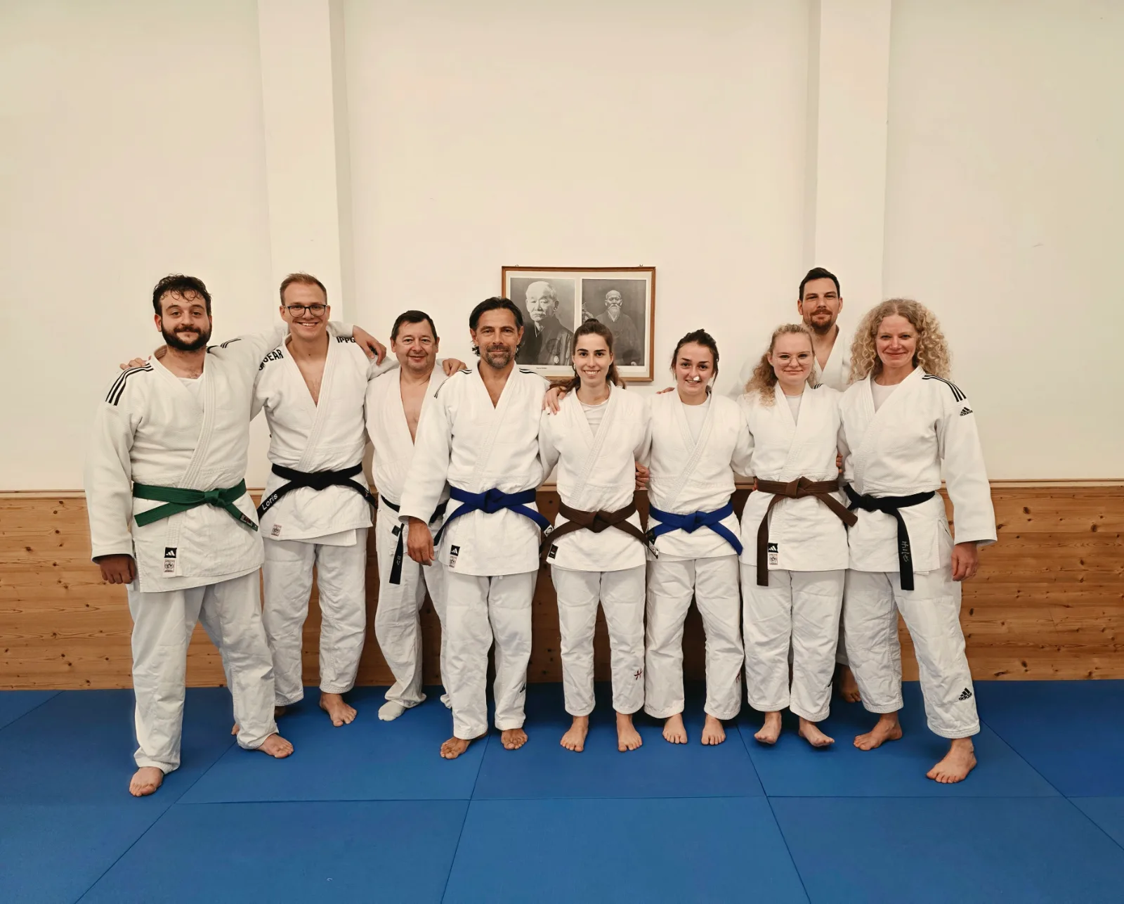 Gruppenfoto von erwachsenen und jugendlichen Judoka in der Turnhalle, alle in Judogi auf der Matte.