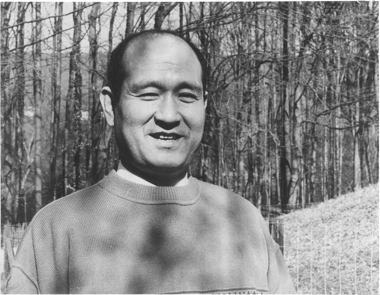 Sensei Masatomi Ikeda, lächelnd im Pullover vor kahlen Bäumen, Schwarz-Weiss-Foto
