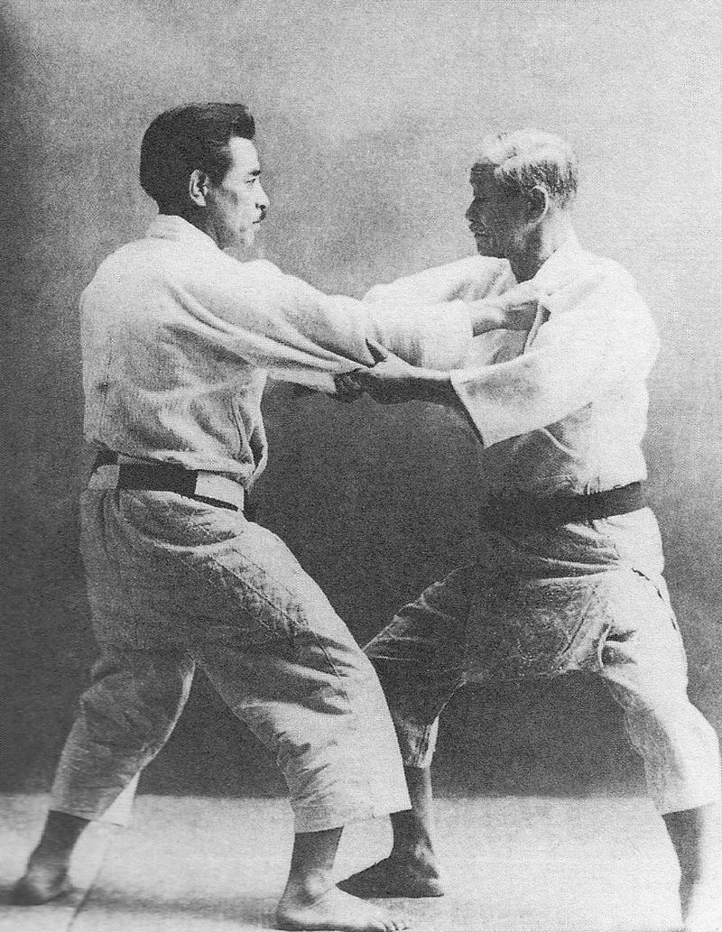 Kanō Jigorō und Kyuzo Mifune in Judogi mit dunklen Gürteln fassen sich an Kragen und Ärmel, Schwarz-Weiss-Foto.