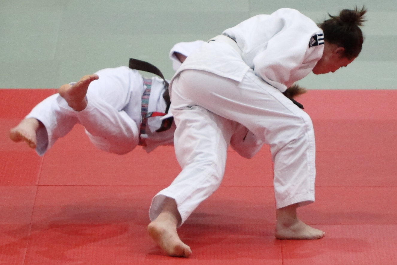 Wettkampf auf roter Matte: Eine Judoka in weissem Judogi wirft ihr Gegenüber, das waagrecht durch die Luft fliegt.
