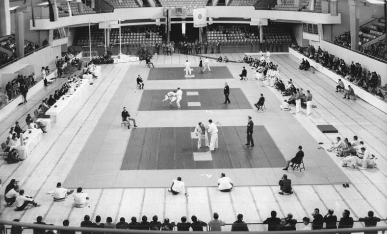 Judo-Wettkampf in einer grossen Halle: Auf drei Mattenflächen kämpfen Paare, daneben Kampfrichter, rundherum Zuschauende, Schwarz-Weiss-Foto.